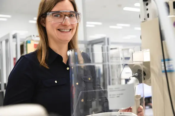 Žena u laboratoriji nosi zaštitne naočale i osmijeh dok stoji pored laboratorijske opreme. U pozadini su regali s knjigama i druge laboratorijske postavke.