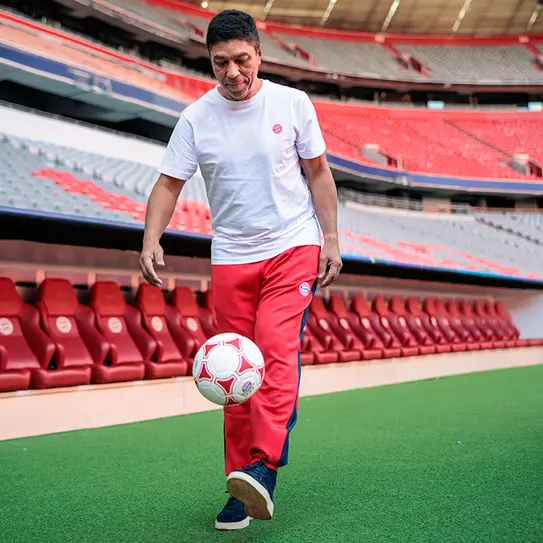 Muškarac u bijeloj majici i crvenim hlačama drži fudbalsku loptu u jednom od svojih ruku, dok se kreće po terenu unutar fudbalskog stadiona s praznim sjedalima u pozadini.