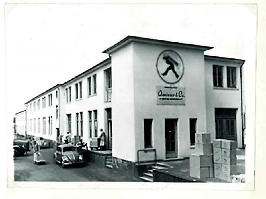 Crno-bijela fotografija zgrade industrijske kompanije s natpisom "Quinver & Co." na fasadi. Ispred zgrade se nalaze neki automobili i ljudi koji rade. Okolo su kutije koje izgledaju kao da su vezane za skladišne aktivnosti.