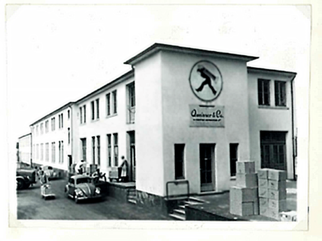 Crno-bijela fotografija zgrade industrijske kompanije s natpisom "Quinver & Co." na fasadi. Ispred zgrade se nalaze neki automobili i ljudi koji rade. Okolo su kutije koje izgledaju kao da su vezane za skladišne aktivnosti.