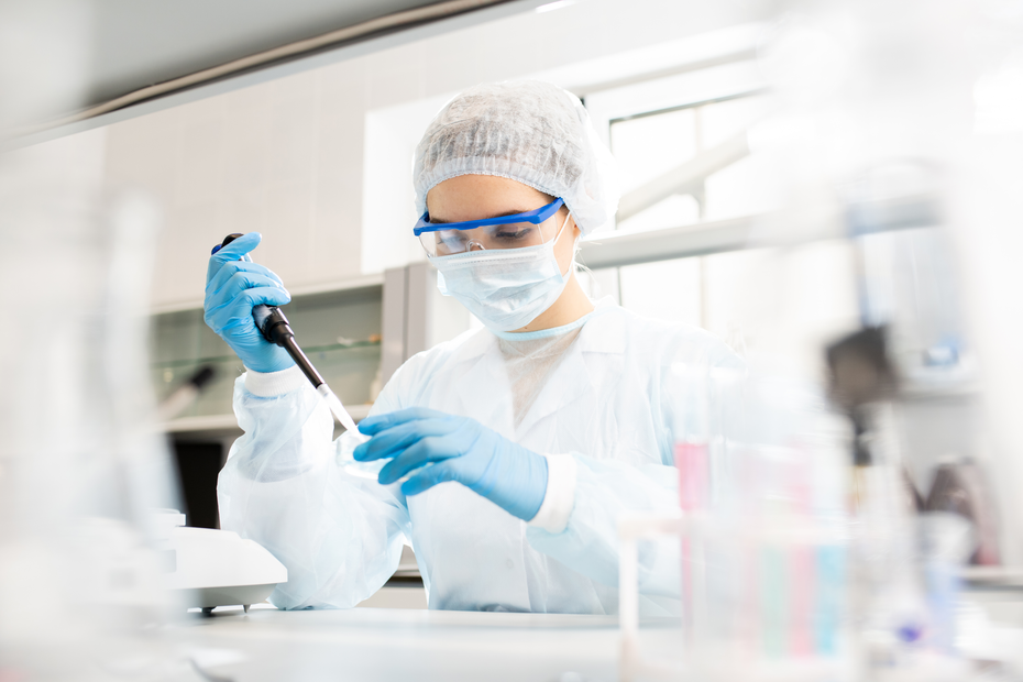 Laborant koristi pipetu u laboratoriji, obučena je u zaštitnu opremu, uključujući masku, rukavice i zaštitne naočare. U pozadini se vide laboratorijski instrumenti i epruvete.