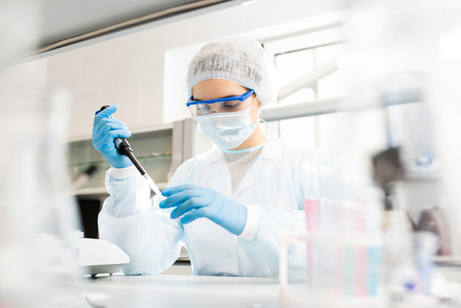 Laborant koristi pipetu u laboratoriji, obučena je u zaštitnu opremu, uključujući masku, rukavice i zaštitne naočare. U pozadini se vide laboratorijski instrumenti i epruvete.