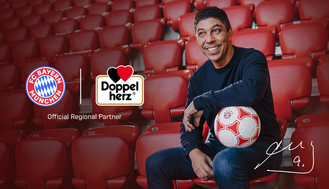 Na slici je nasmijani muškarac sjedi na stadionu sa crvenim sjedalima, držeći bijelu fudbalsku loptu. Iza njega su logotipi FC Bayerna, Doppelherza i oznaka "Official Regional Partner". Atmosfera je prijateljska i sportska.