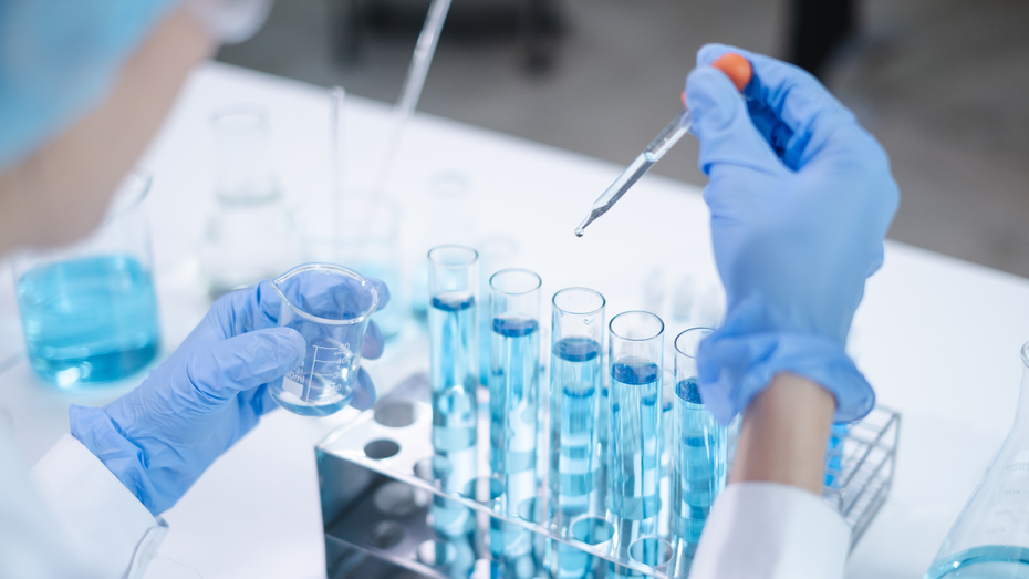Osoba u laboratorijskim rukavicama drži mjernu posudu i pipetu dok dodaje plavu tečnost u niz epruveta. U pozadini se vide i druge laboratorijske boce s plavom tečnošću, stvarajući naučnu atmosferu.