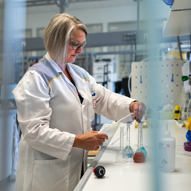 Žena u laboratorijskom kutu miješa tekućine u labaratoriji. Oko nje su različite staklene boce i posude, a ona pažljivo koristi pipetu. U pozadini se vide drugi laboratorijski uređaji i oprema.