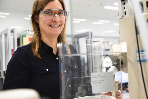 Žena u laboratoriji nosi zaštitne naočale i osmijeh dok stoji pored laboratorijske opreme. U pozadini su regali s knjigama i druge laboratorijske postavke.
